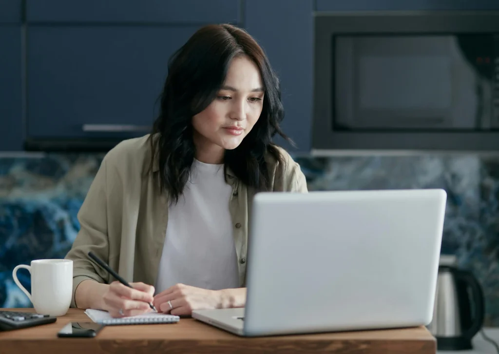 woman writing in notebook while looking at open laptop in front of her